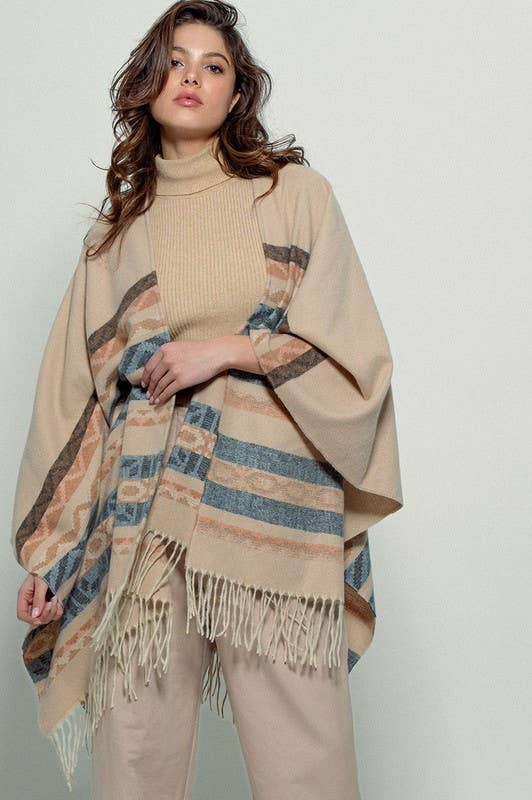 Woman wearing a beige poncho with a Tan background and light blue, brown, light rust, and grey aztec pattern design in stripes with tassel trim along bottom hem. Light weight fabric