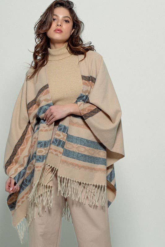 Woman wearing a beige poncho with a Tan background and light blue, brown, light rust, and grey aztec pattern design in stripes with tassel trim along bottom hem. Light weight fabric