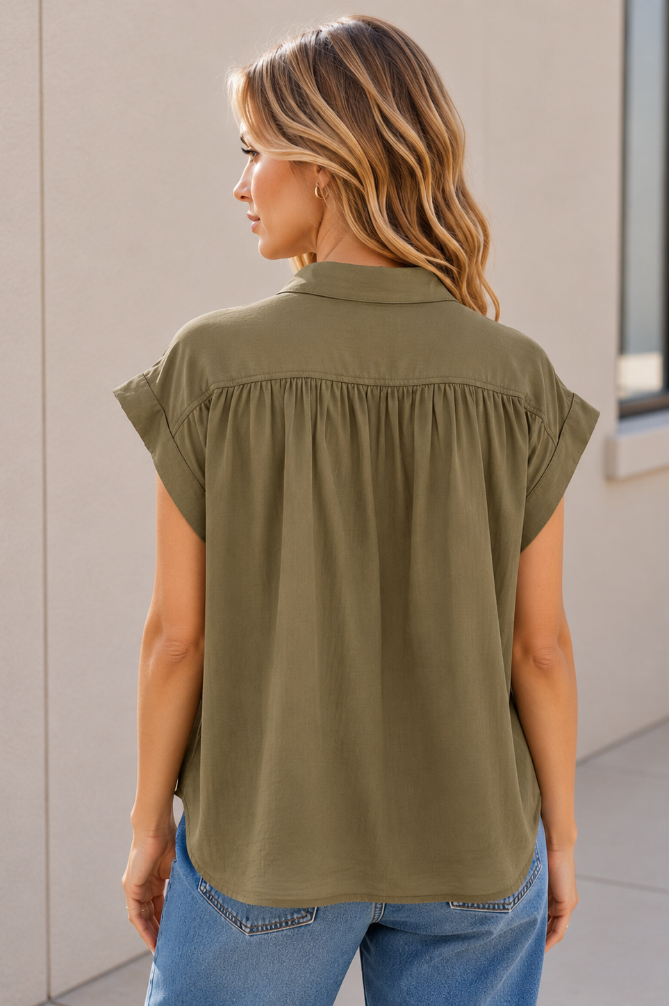 A woman shown from the back wearing an olive green /khaki short-sleeve blouse with a gathered back seam, paired with blue jeans, standing against a light beige wall.