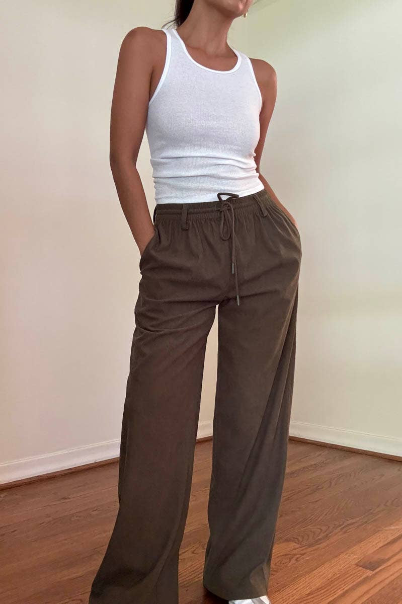 Person wearing a white tank top and brown high-waisted pants in a minimal setting.