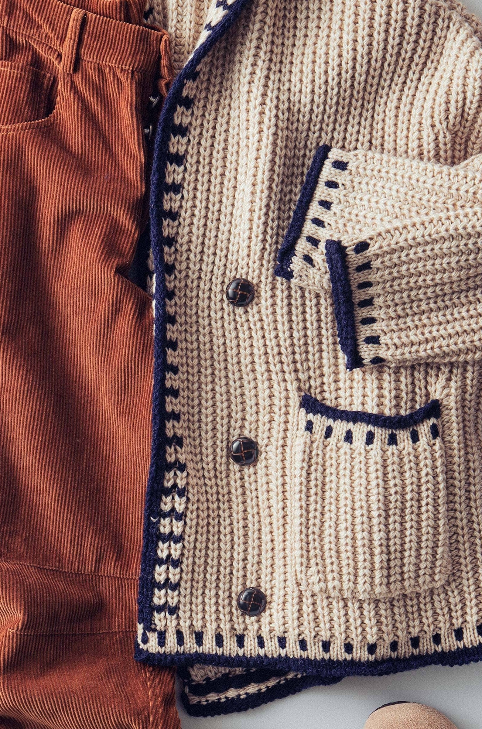 Close-up of a beige knitted cardigan with navy trim and buttons, paired with brown pants.