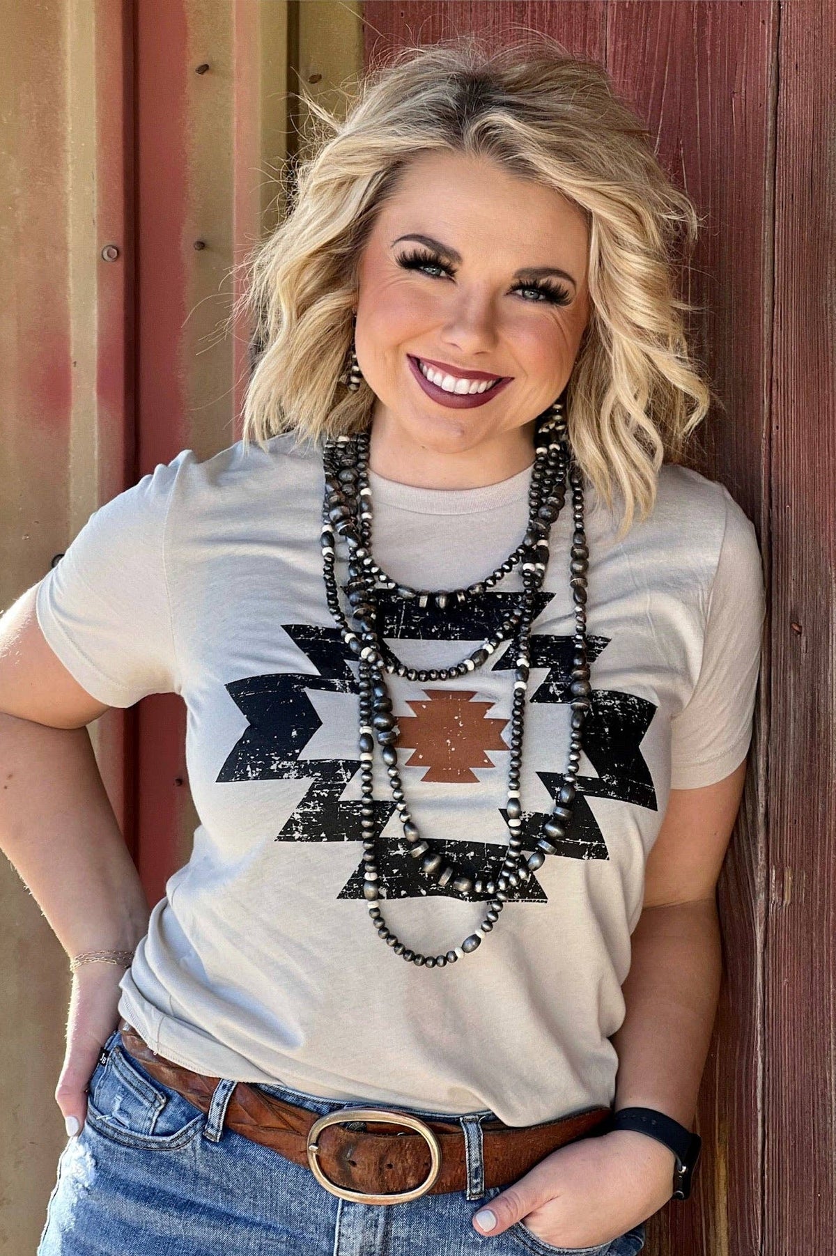 Woman wearing a white t-shirt with black geometric patterns, standing against a wooden wall.