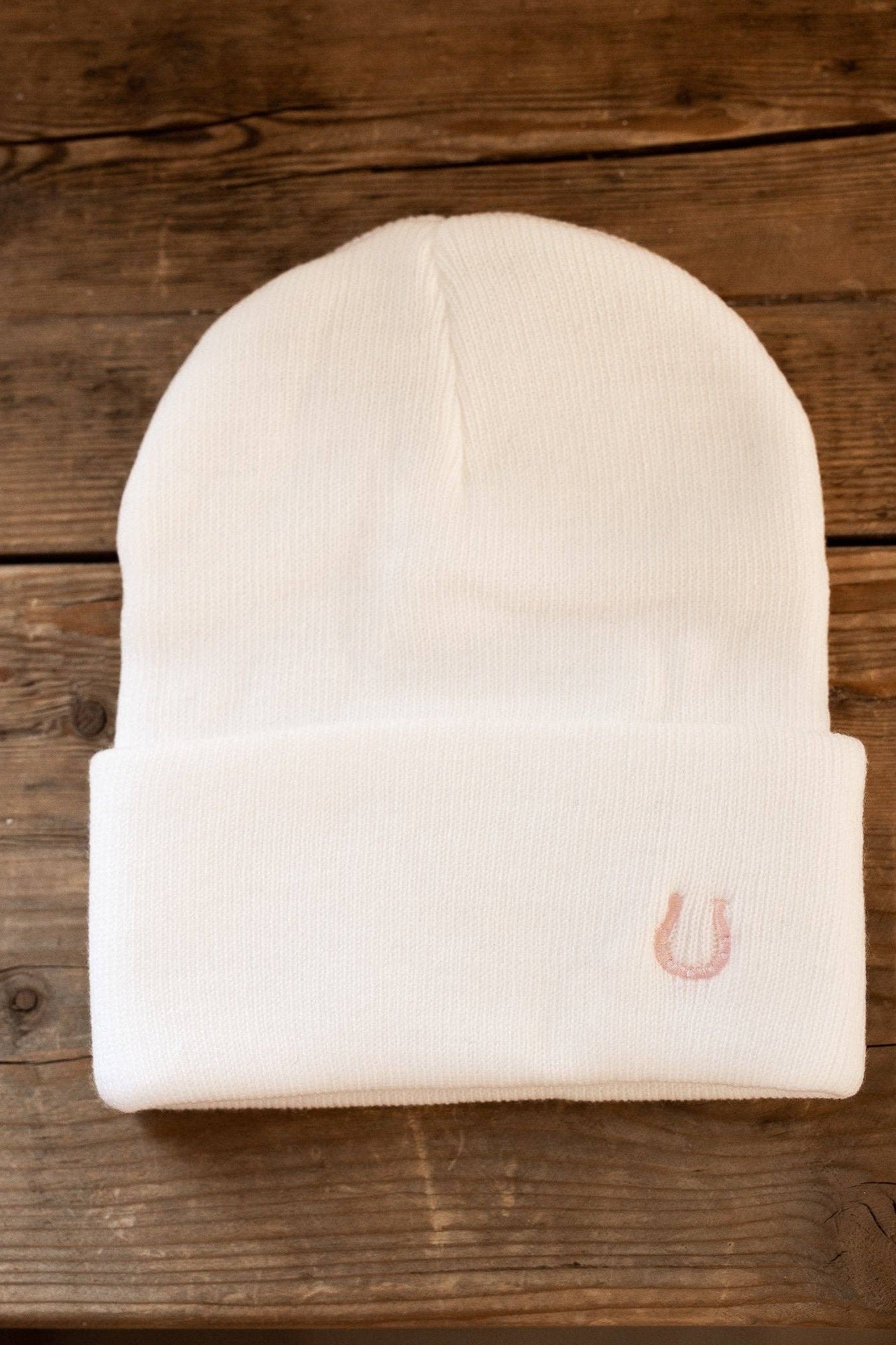 White beanie with a embroidered small pink horseshoe on beanie cuff on a wooden surface