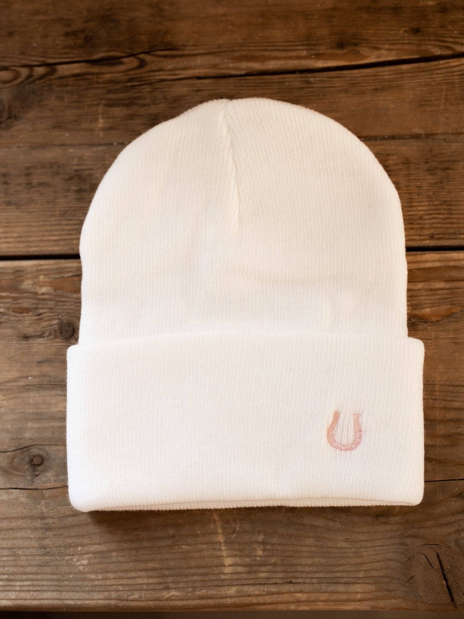 White beanie with a embroidered small pink horseshoe on beanie cuff on a wooden surface