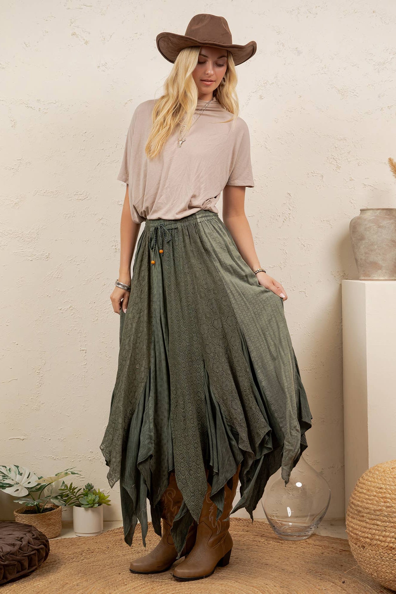 A woman stands in a styled indoor setting wearing a brown wide-brim hat, a light beige short-sleeve top, a long olive green tiered skirt, and brown ankle boots. The room has a textured cream wall, woven poufs, plants, and decorative vases.