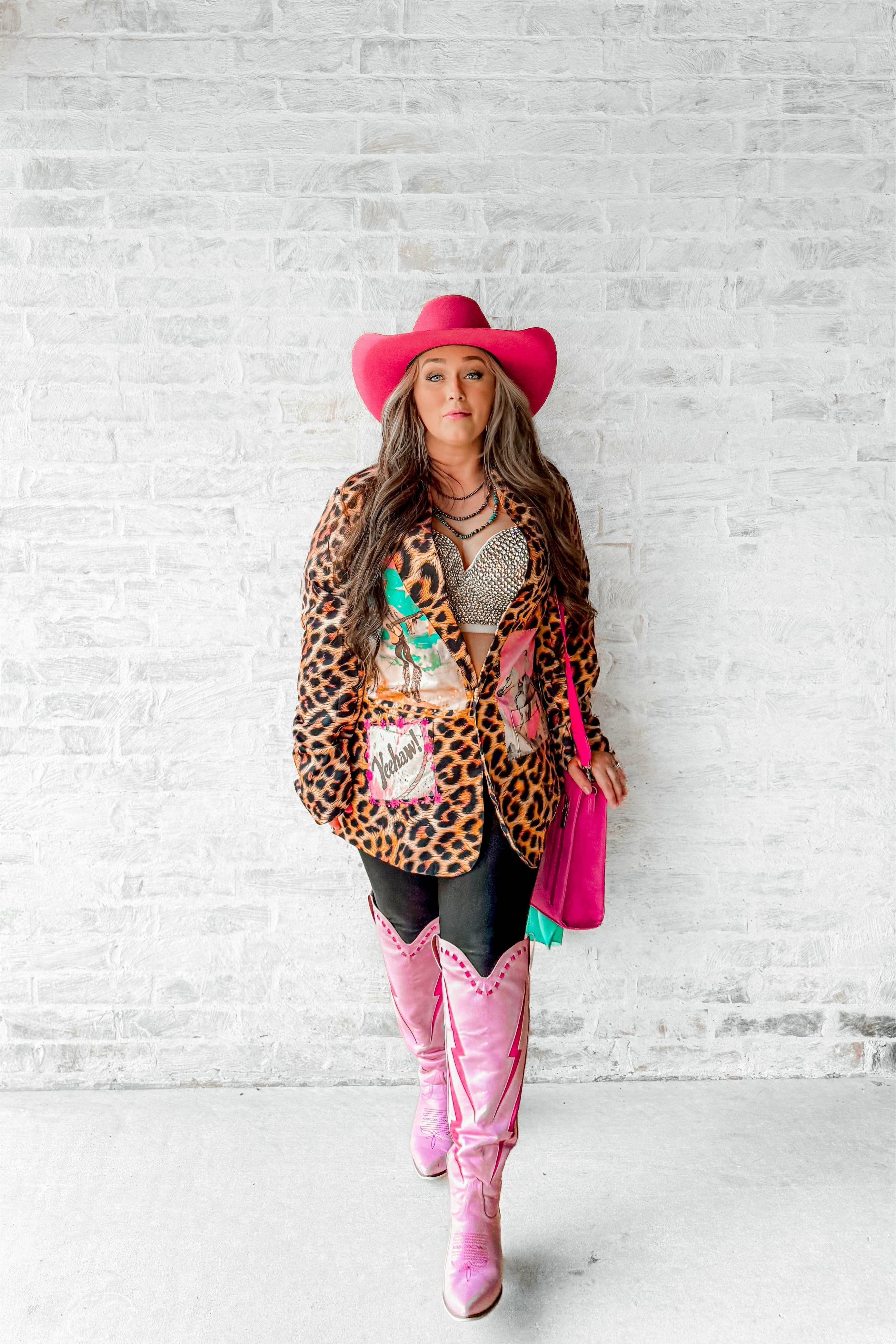 Person wearing a leopard print jacket, pink cowboy hat, and pink boots against a white brick wall.