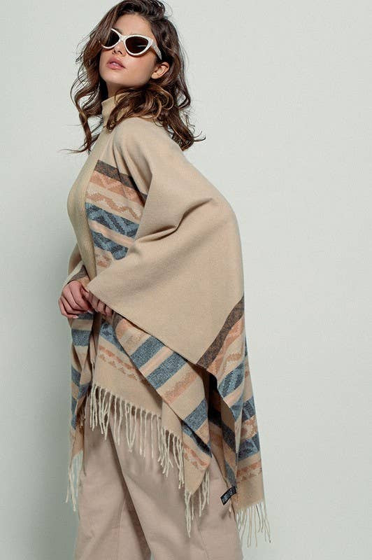 Woman wearing a beige poncho with a Tan background and light blue, brown, light rust, and grey aztec pattern design in stripes with tassel trim along bottom hem. Light weight fabric