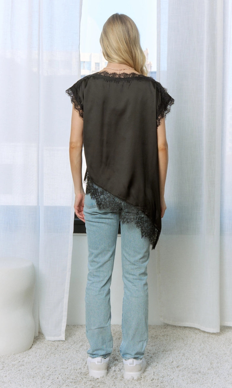 Back View: Black satin asymmetric top with lace-trim neckline, short sleeves, and diagonal lace hem, styled with light-wash jeans.