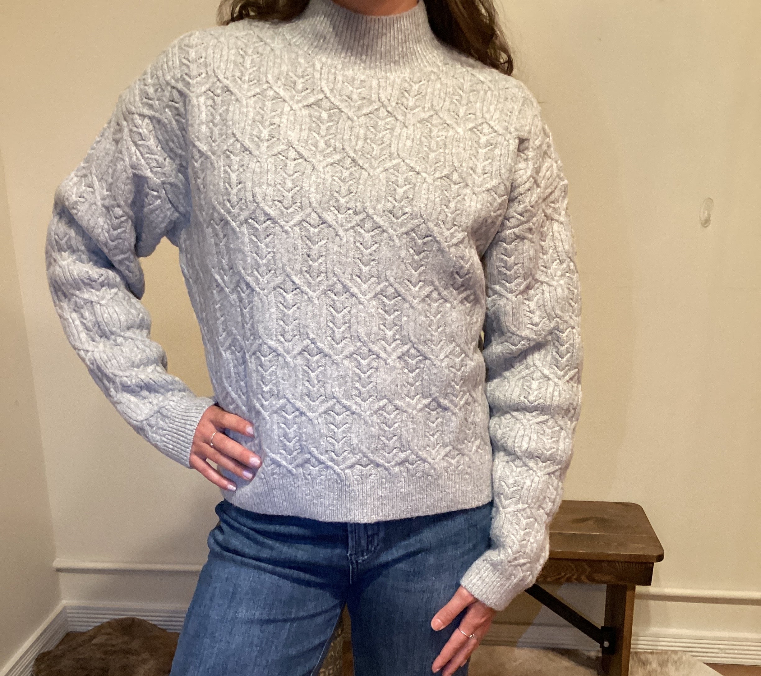 Person wearing a gray cable knit sweater and blue jeans standing against a neutral wall.