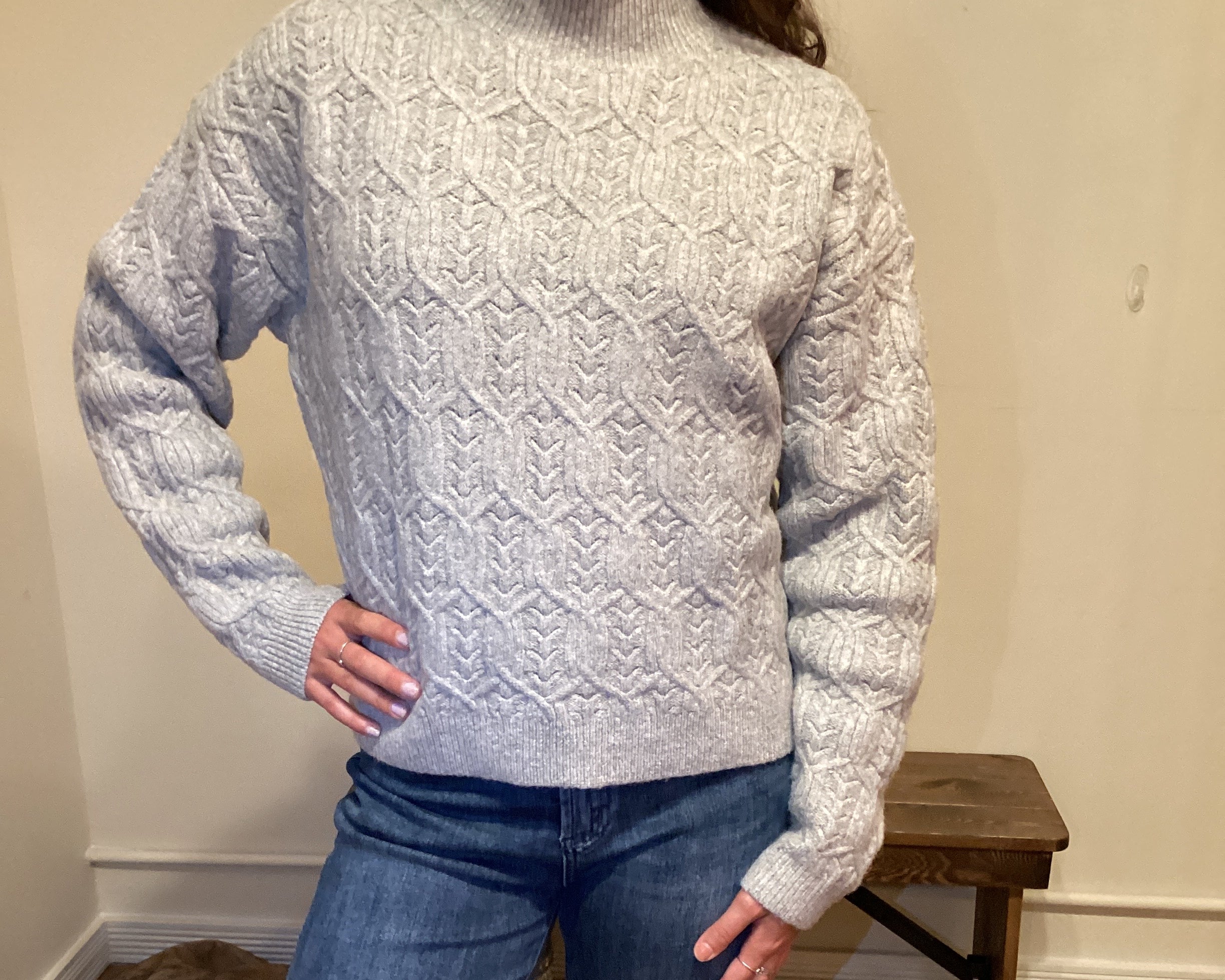 Person wearing a gray cable knit sweater and blue jeans standing against a neutral wall.