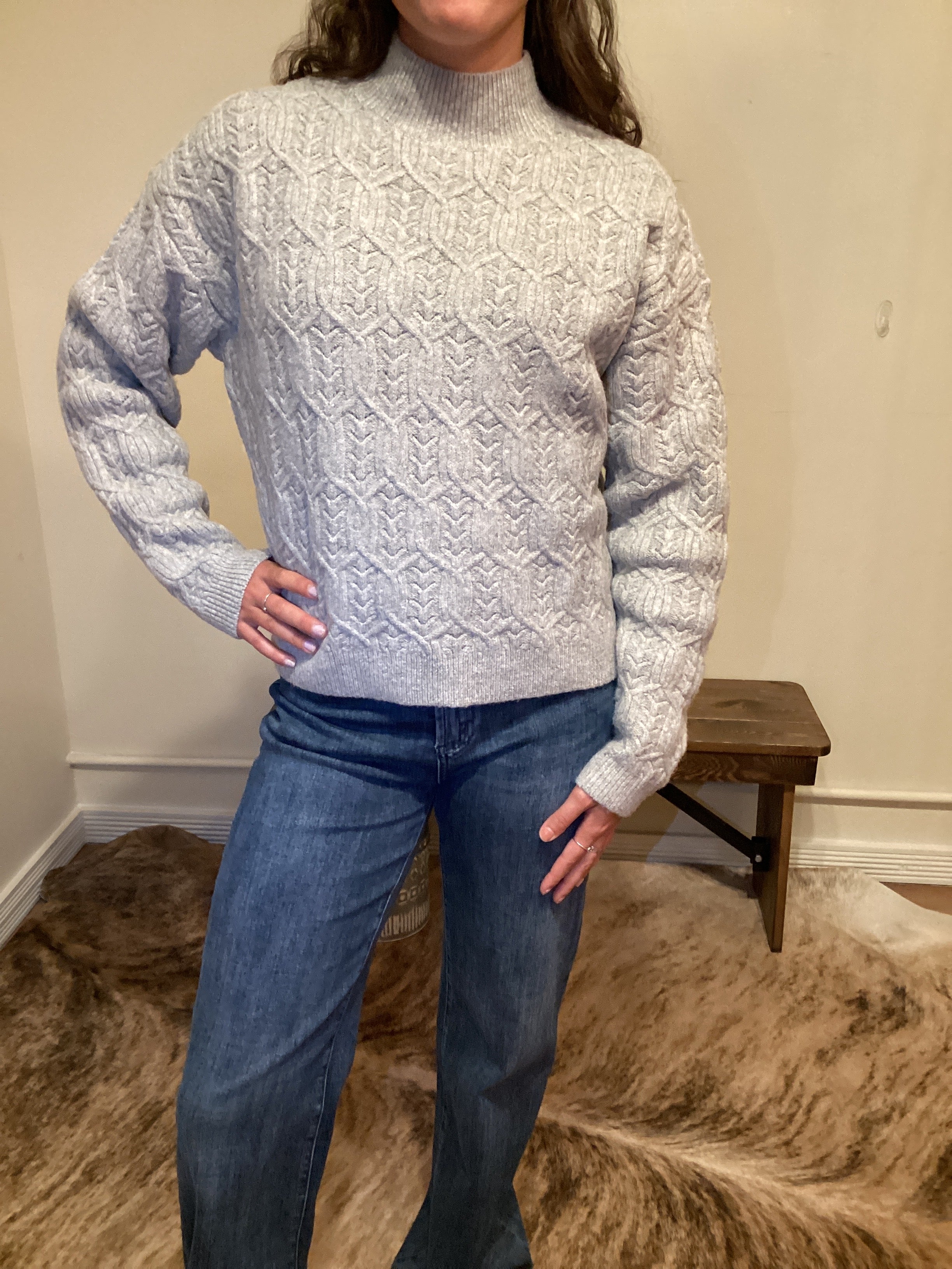 Person wearing a gray cable knit sweater and blue jeans standing against a neutral wall.