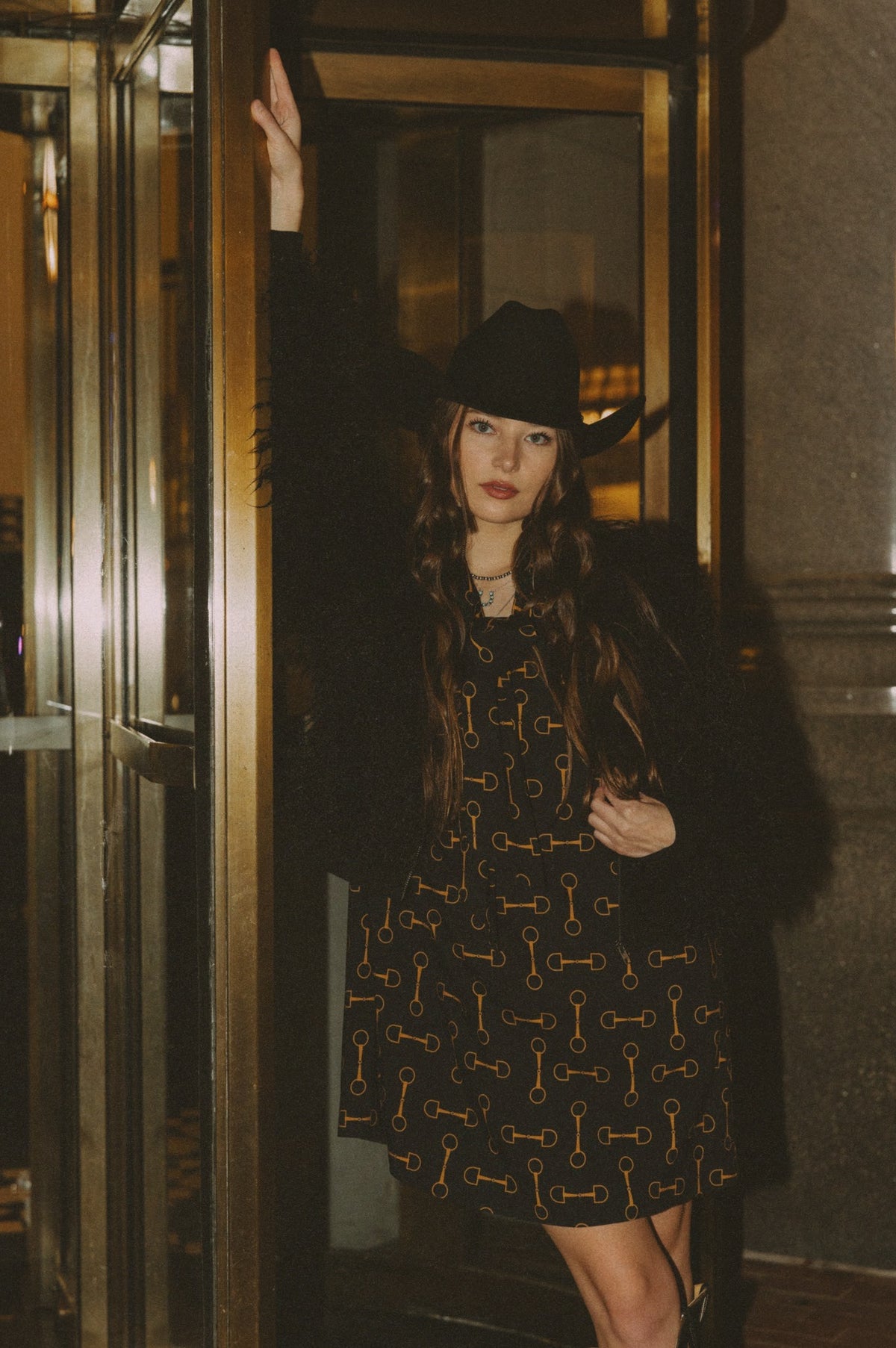 Woman in a patterned dress and fur coat standing in front of a mirror.