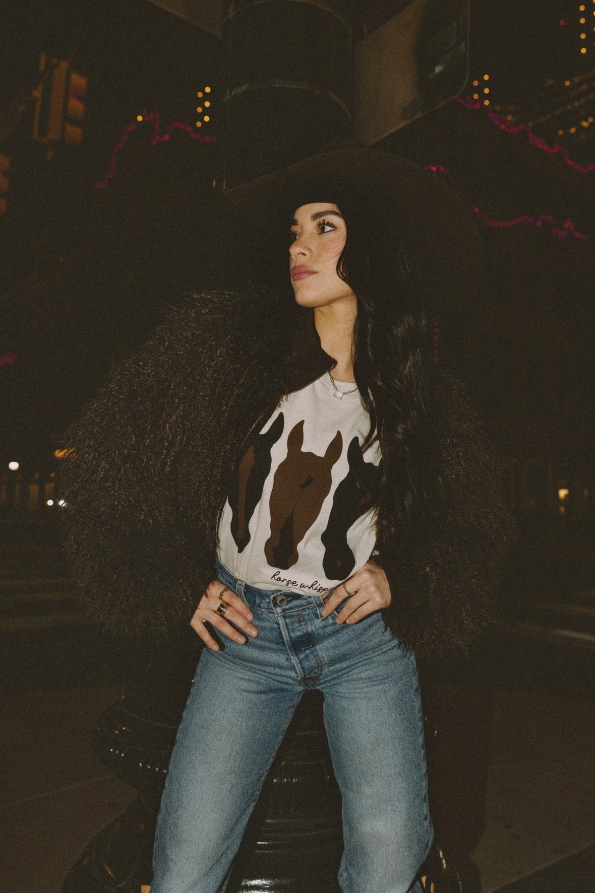 Woman wearing a white sweater with a horse design, brown faux fur jacket and blue jeans at night.