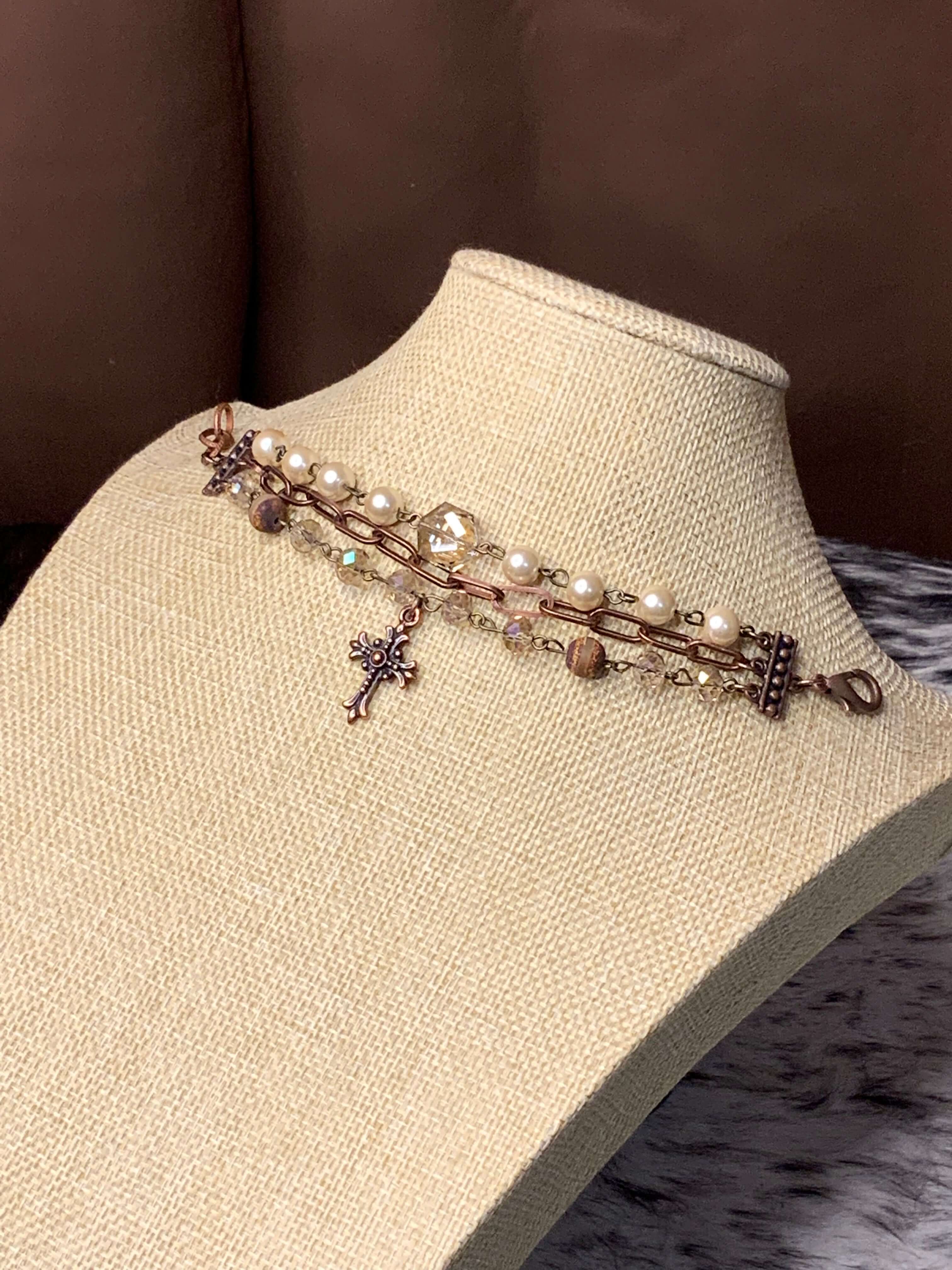 A handcrafted bracelet featuring pearls, crystals, a copper cross charm, and agate stone beads, displayed on a mannequin bust.