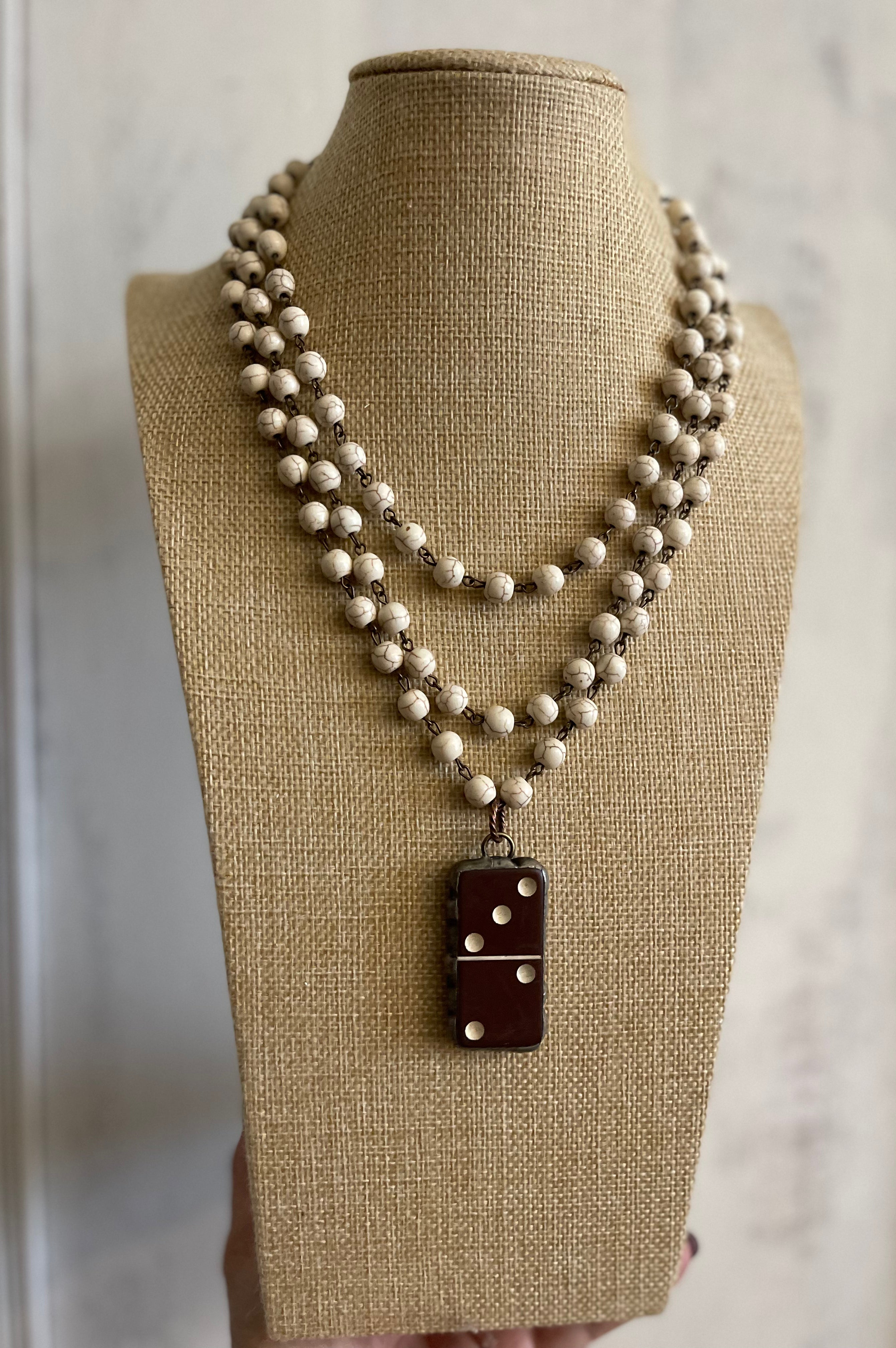 A multi-strand beaded necklace displayed on a burlap-covered mannequin bust. The necklace features cream-colored beads with small metallic spacer beads, and a dark reddish-brown rectangular pendant with white dot details.