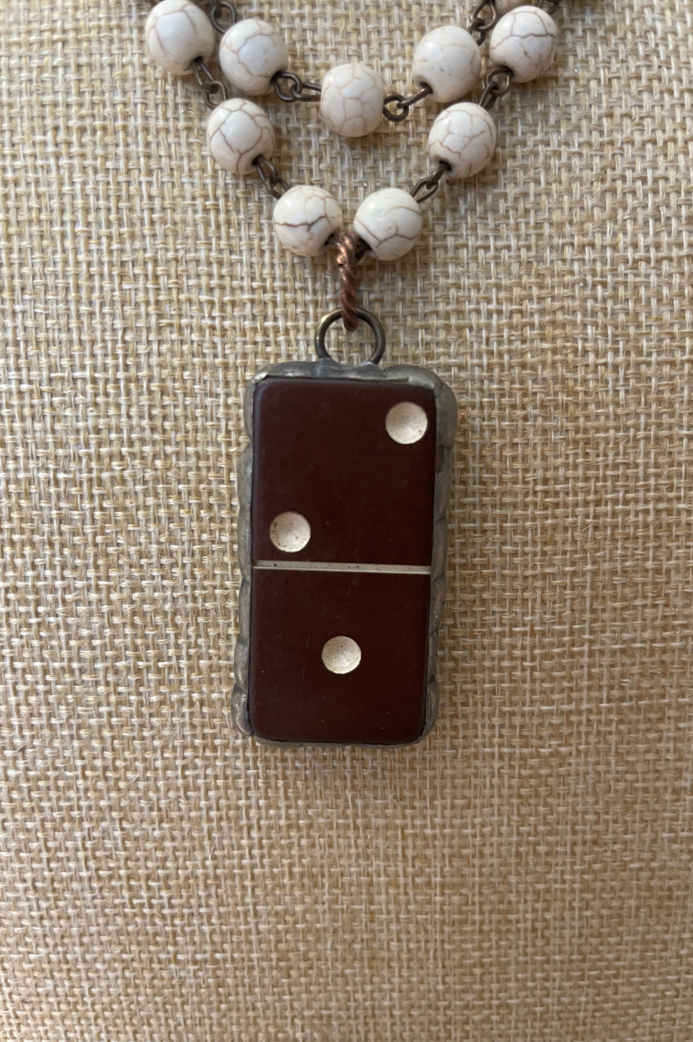 A close-up of a necklace with a dark brown domino-shaped pendant and white marbled beads on a chain, displayed on a beige burlap background.