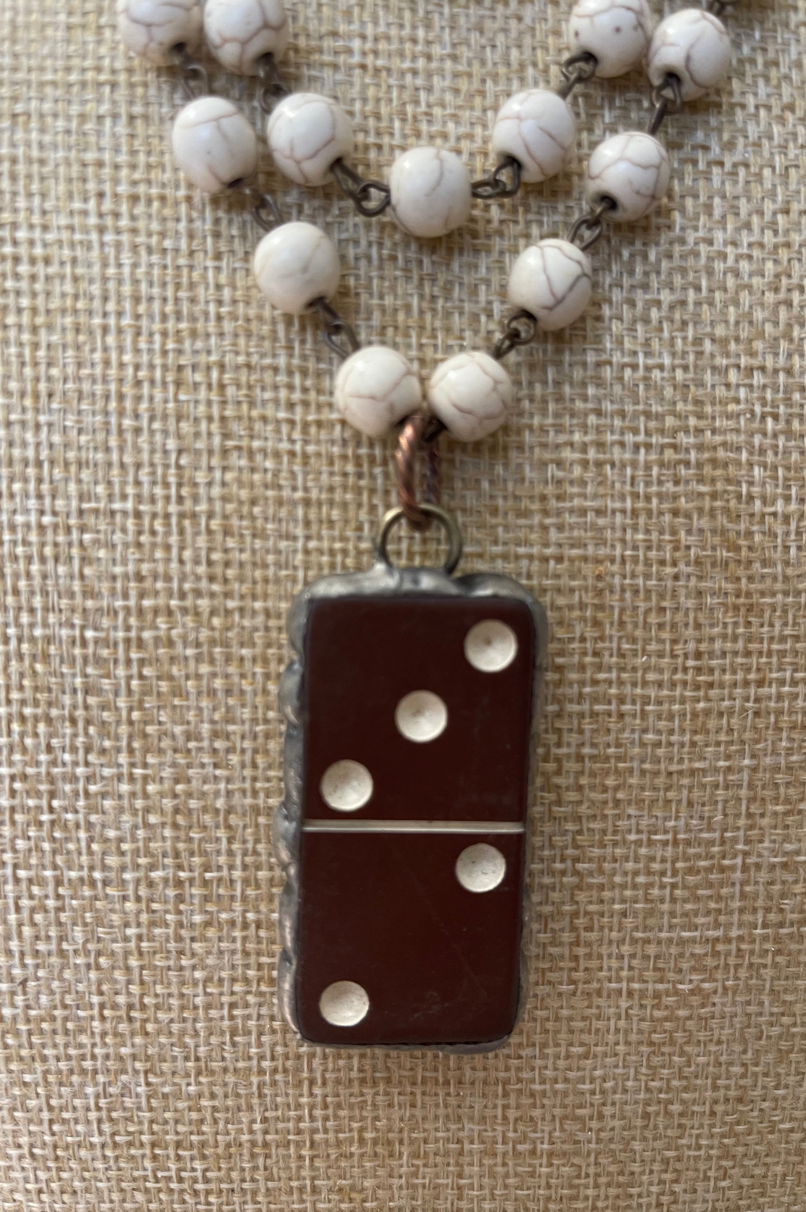 A close-up of a necklace with a dark brown domino-shaped pendant and white marbled beads on a textured beige fabric background.