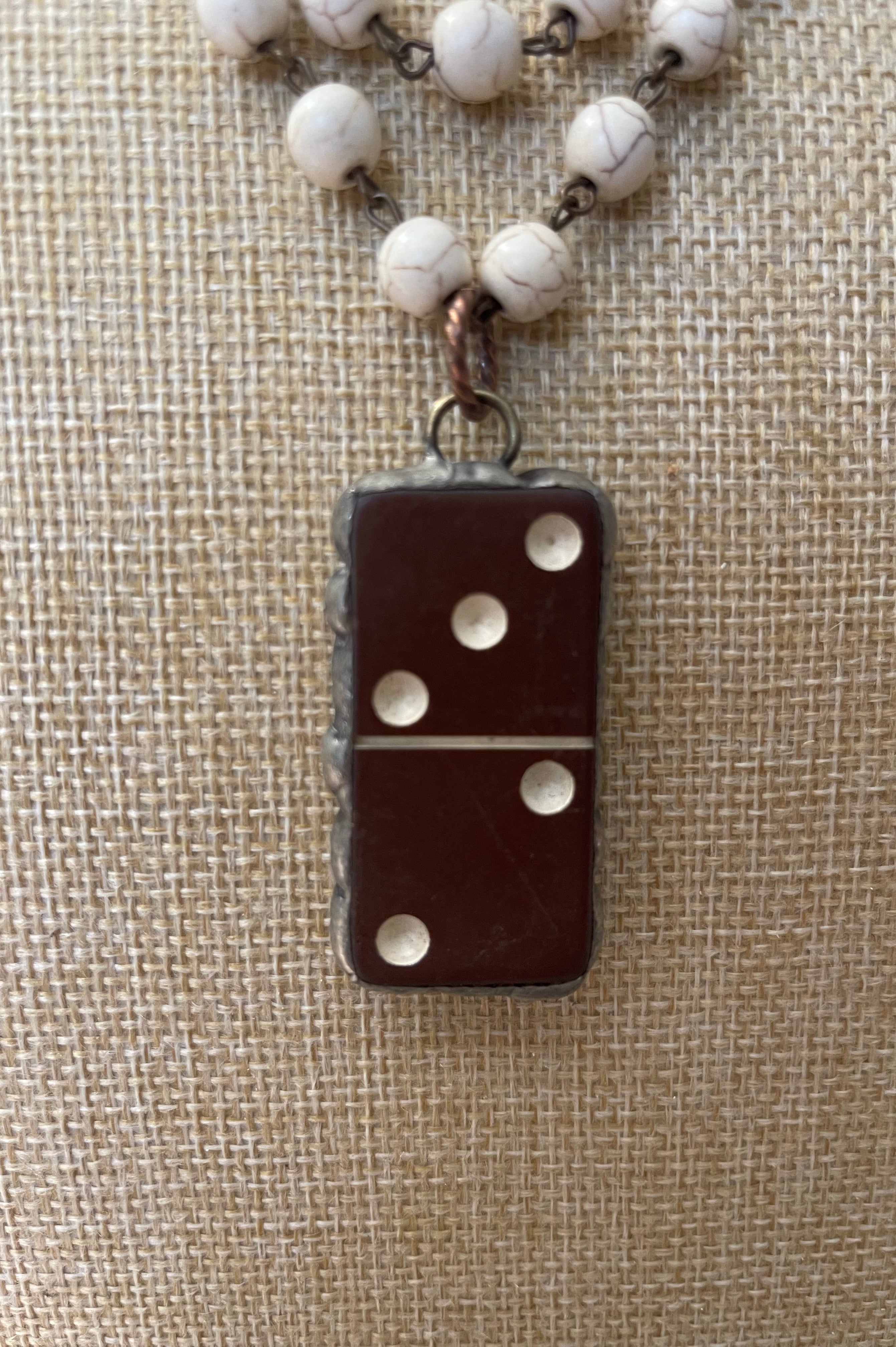 A close-up of a necklace with a dark brown domino-shaped pendant and white marbled beads on a chain, displayed on a beige burlap background.