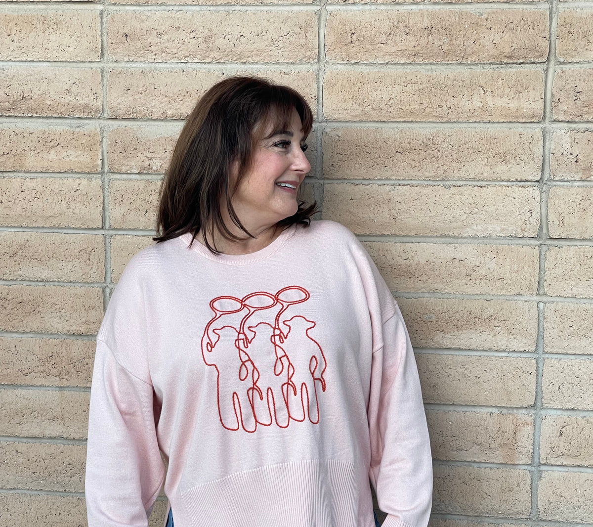 A person wearing a light pink crewneck sweatshirt with a orange line-art embroidery graphic of three cowboy figures, paired with blue jeans, standing against a light tan brick wall.