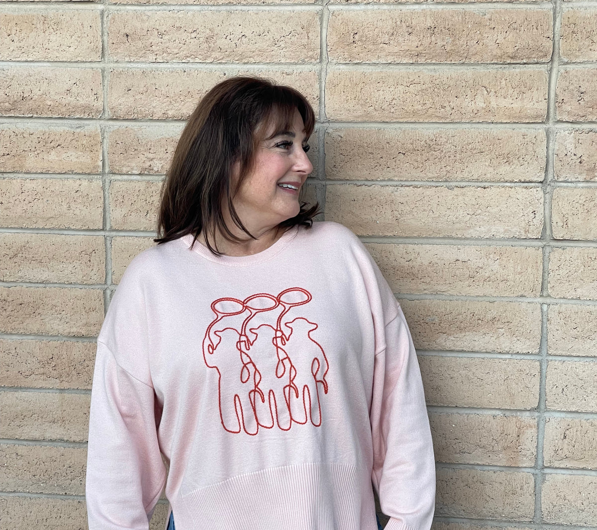 A person wearing a light pink crewneck sweatshirt with a orange line-art embroidery graphic of three cowboy figures, paired with blue jeans, standing against a light tan brick wall.