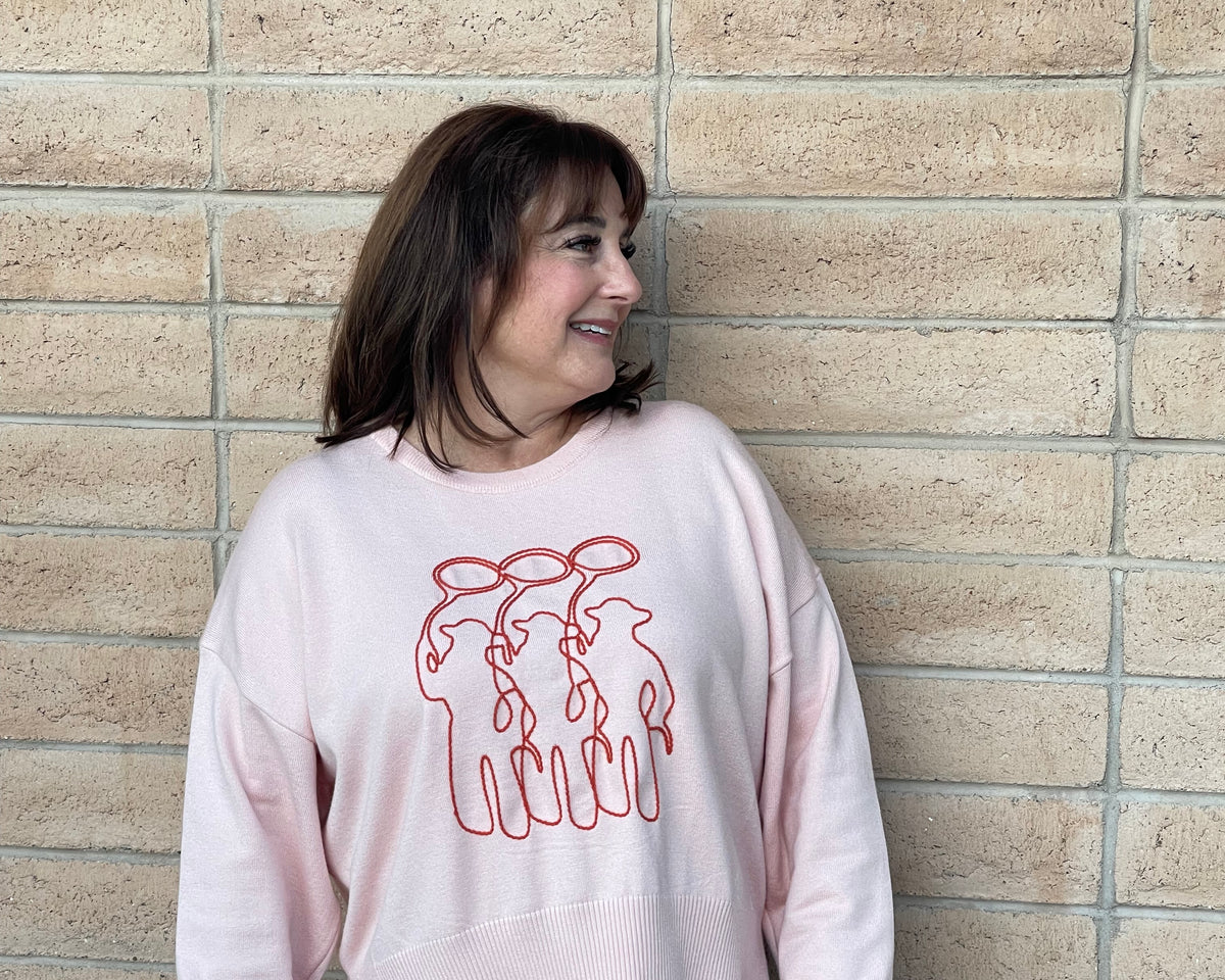 A person wearing a light pink crewneck sweatshirt with a orange line-art embroidery graphic of three cowboy figures, paired with blue jeans, standing against a light tan brick wall.