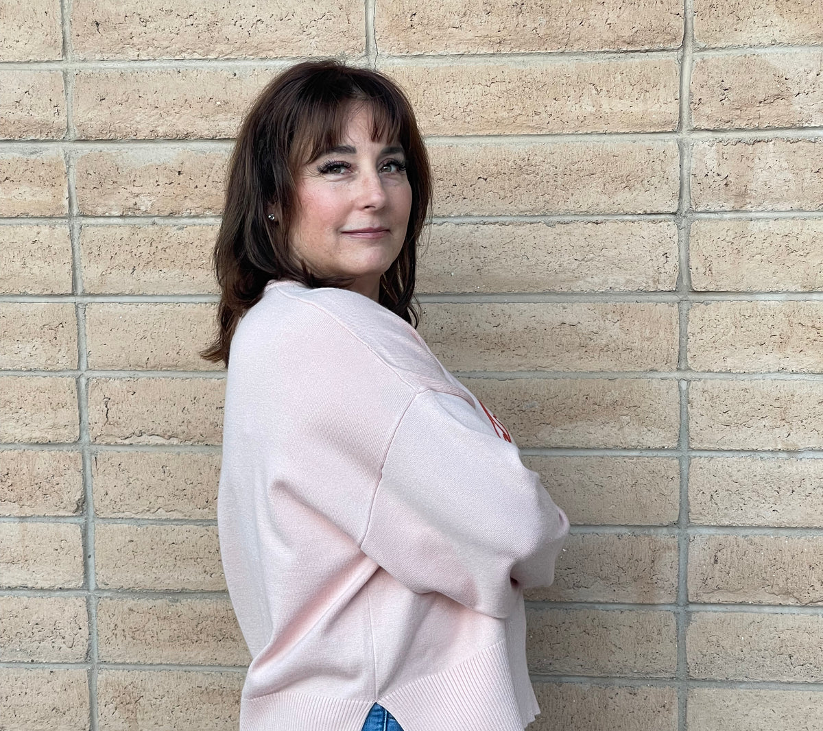 A woman with brown hair stands against a beige brick wall, wearing a light pink ribbed knit sweater and blue jeans.