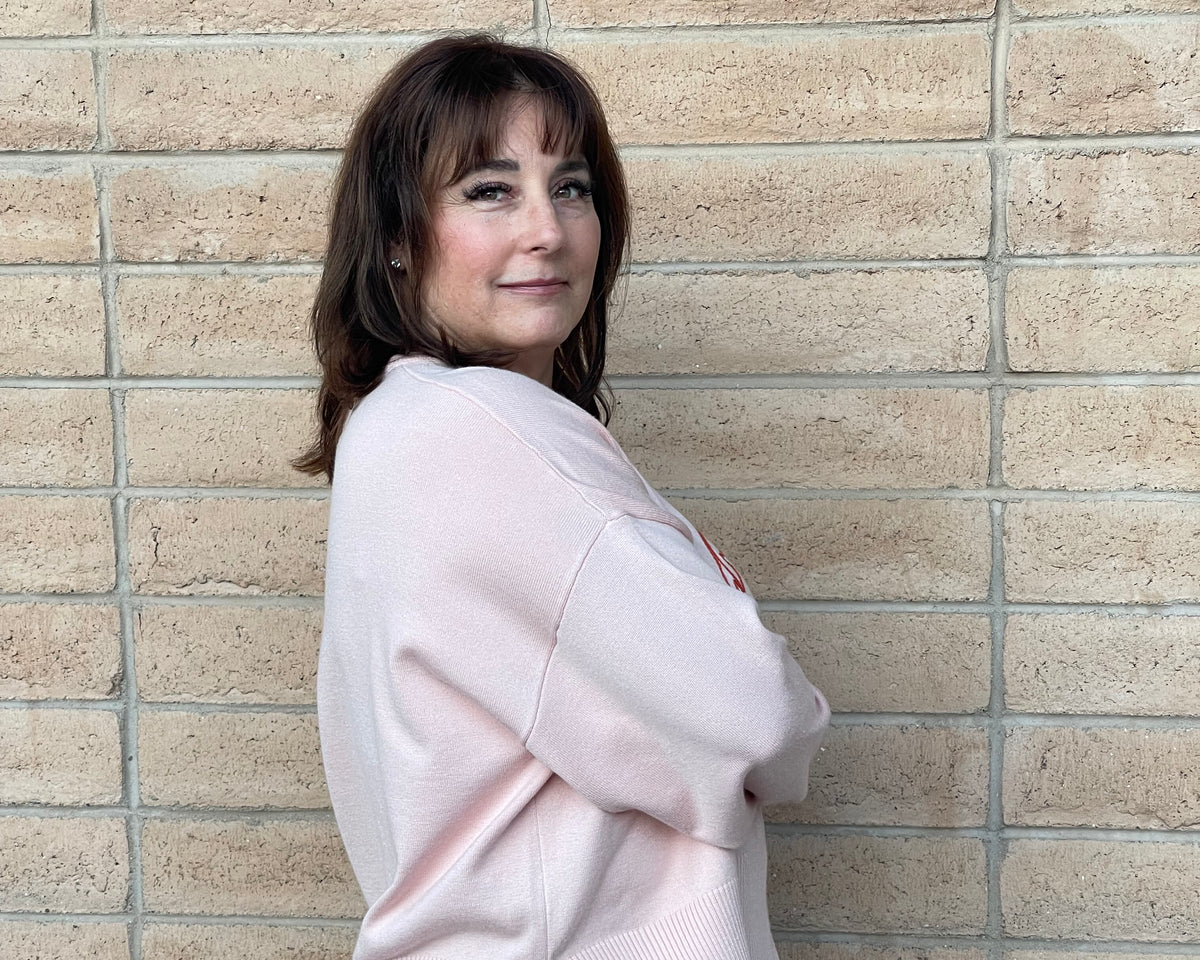 A woman with brown hair stands against a beige brick wall, wearing a light pink ribbed knit sweater and blue jeans.