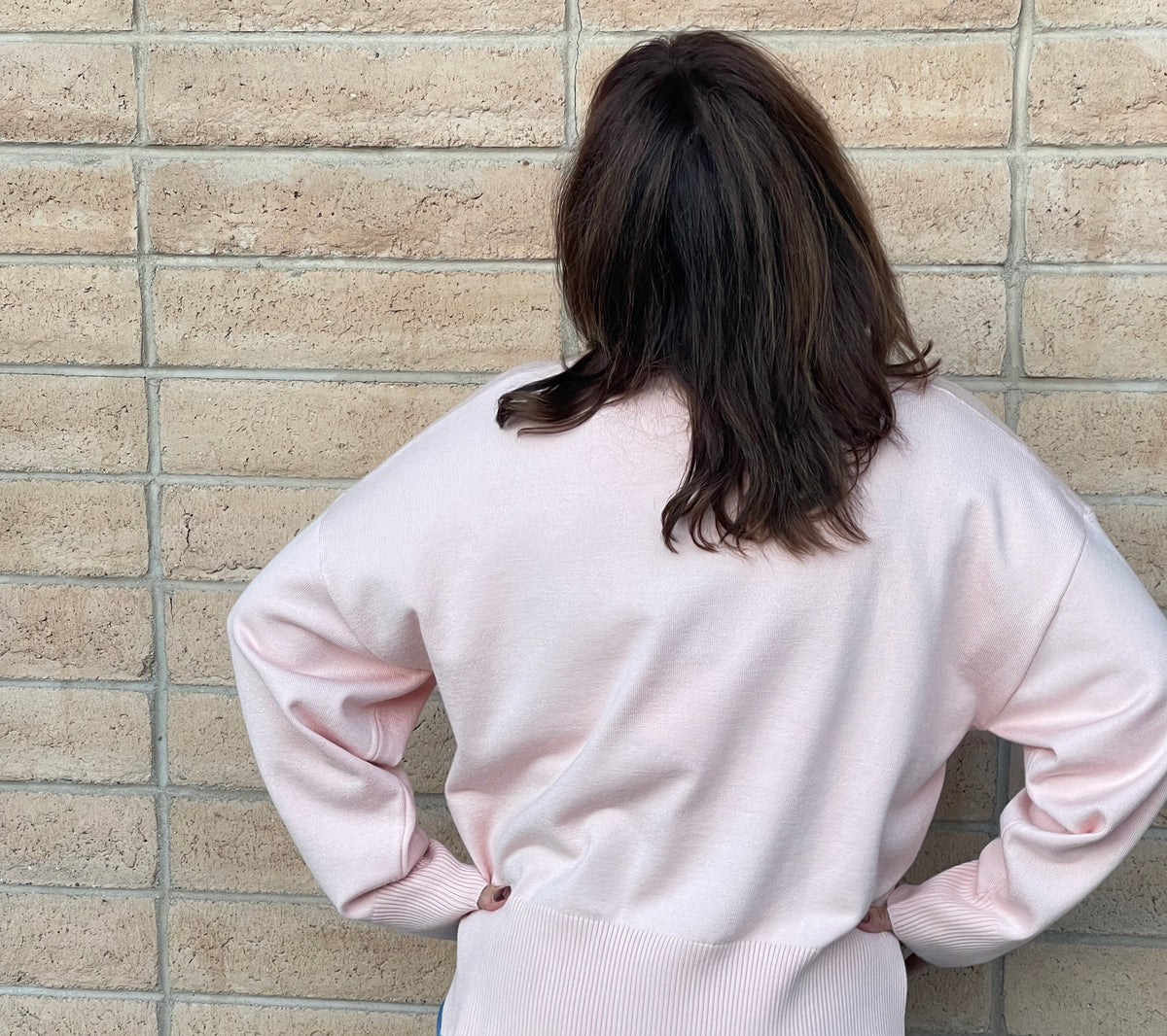 A person viewed from behind wearing a light pink knit sweater and blue jeans, standing against a beige brick wall.
