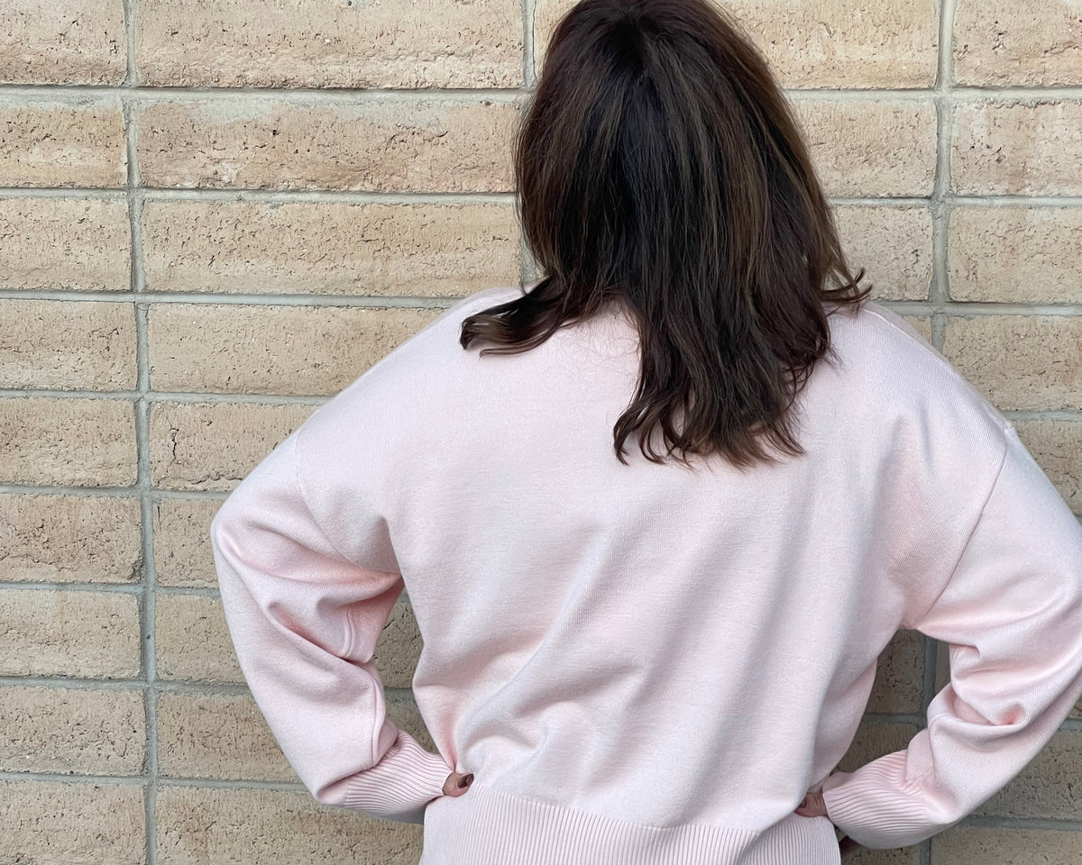 A person viewed from behind wearing a light pink knit sweater and blue jeans, standing against a beige brick wall.