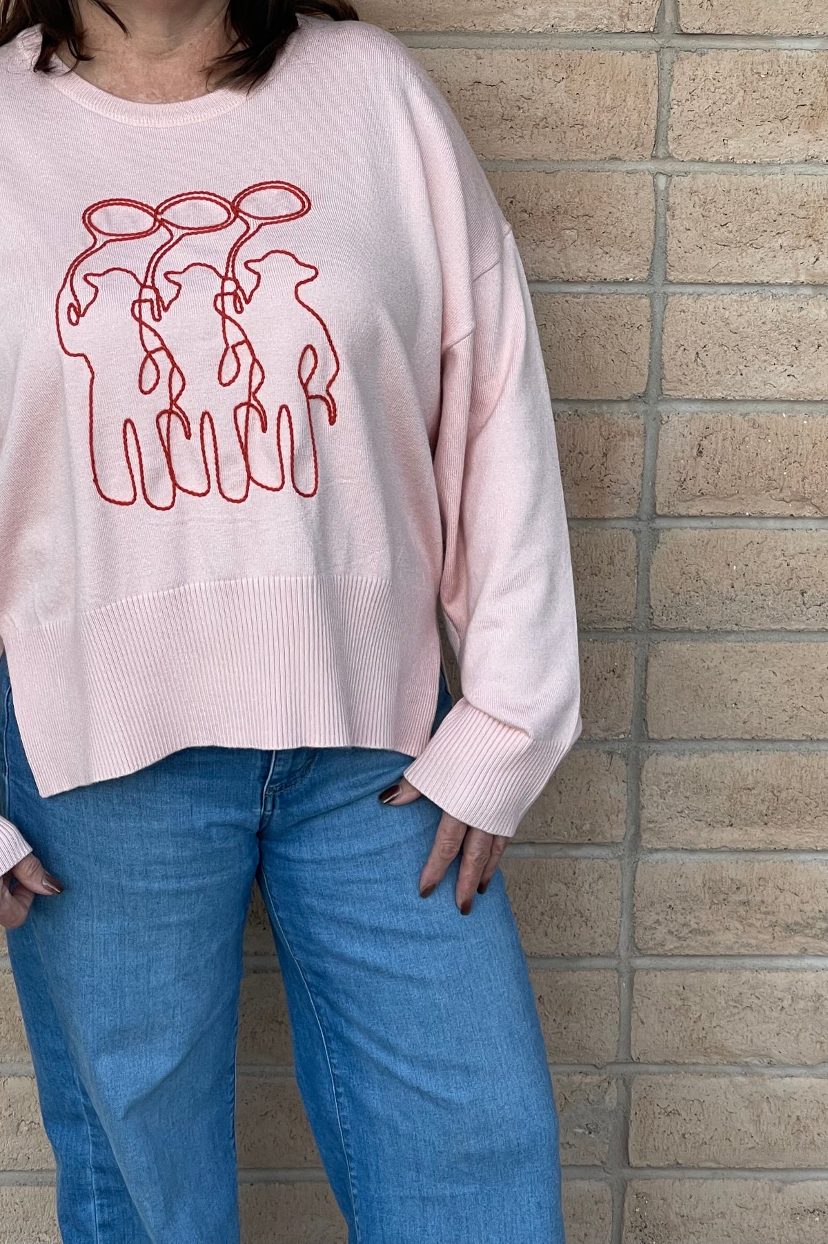 A person wearing a light pink crewneck sweatshirt with a orange line-art embroidery graphic of three cowboy figures, paired with blue jeans, standing against a light tan brick wall.