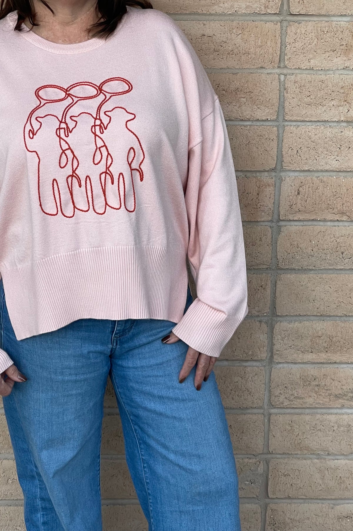 A person wearing a light pink crewneck sweatshirt with a orange line-art embroidery graphic of three cowboy figures, paired with blue jeans, standing against a light tan brick wall.