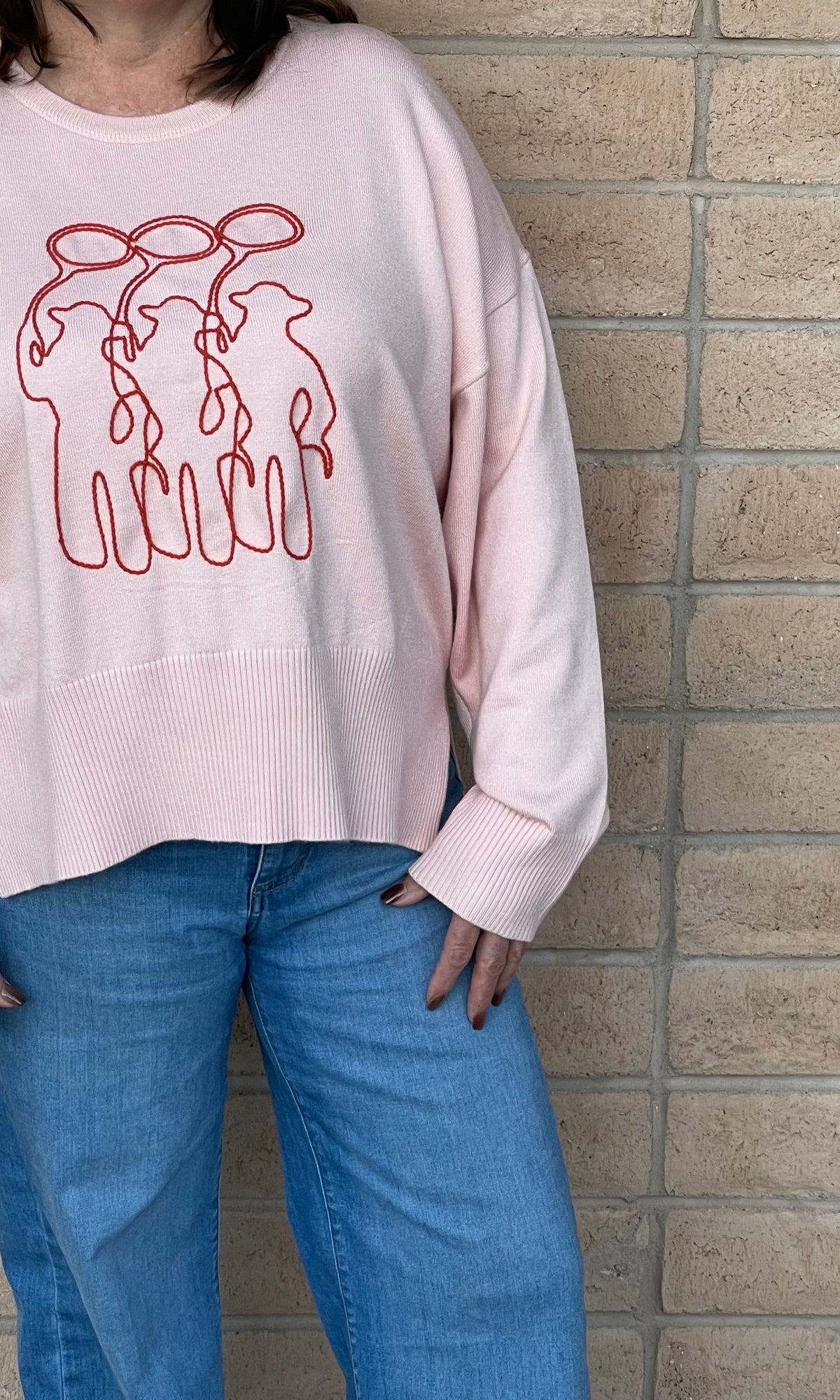 A person wearing a light pink crewneck sweatshirt with a orange line-art embroidery graphic of three cowboy figures, paired with blue jeans, standing against a light tan brick wall.