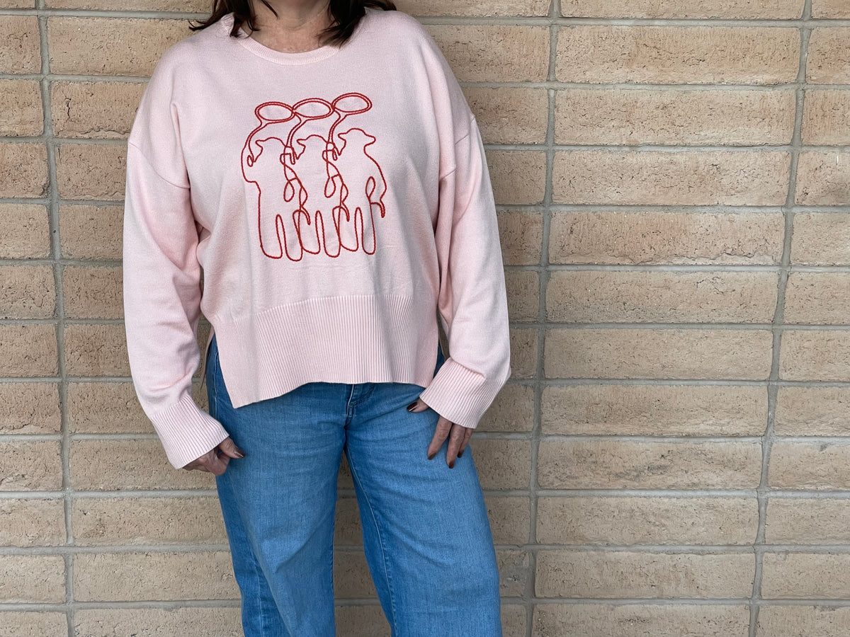 A person wearing a light pink crewneck sweatshirt with a orange line-art embroidery graphic of three cowboy figures, paired with blue jeans, standing against a light tan brick wall.