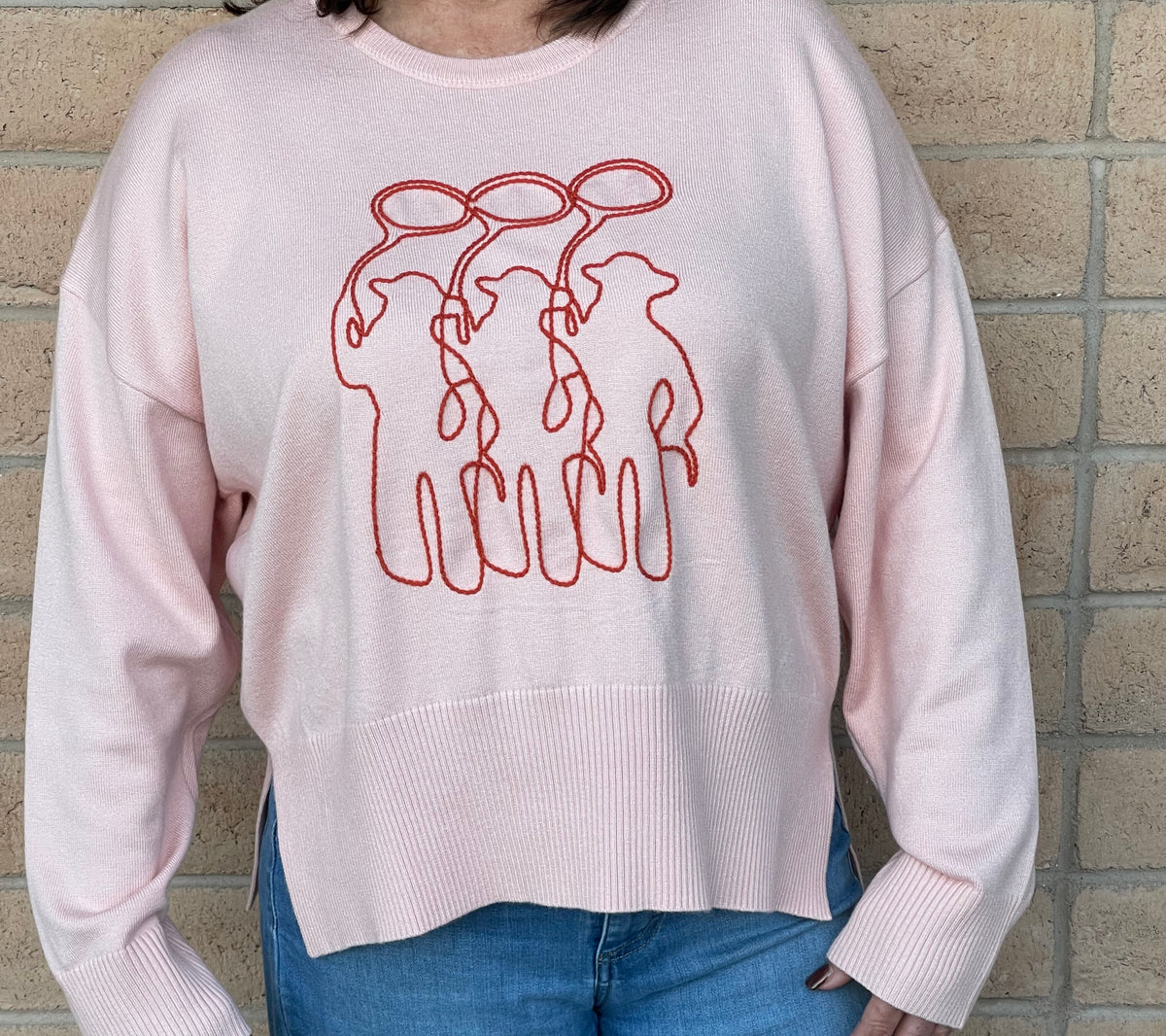 A person wearing a light pink crewneck sweatshirt with a orange line-art embroidery graphic of three cowboy figures, paired with blue jeans, standing against a light tan brick wall.