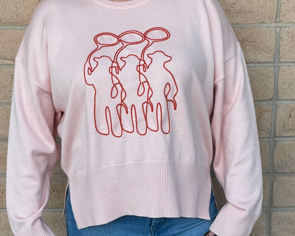 A person wearing a light pink crewneck sweatshirt with a orange line-art embroidery graphic of three cowboy figures, paired with blue jeans, standing against a light tan brick wall.