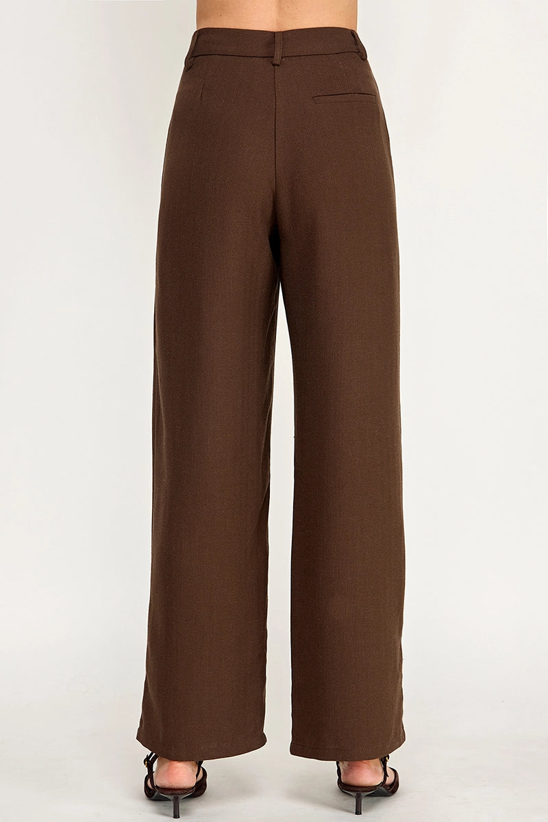 A person shown from the back wearing high-waisted, wide-leg brown trousers and black heeled sandals against a plain light gray studio background.