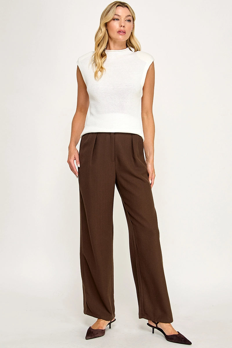 A woman modeling a white sleeveless mock-neck knit top and dark brown wide-leg trousers, paired with dark brown pointed-toe heels, against a plain white studio background.