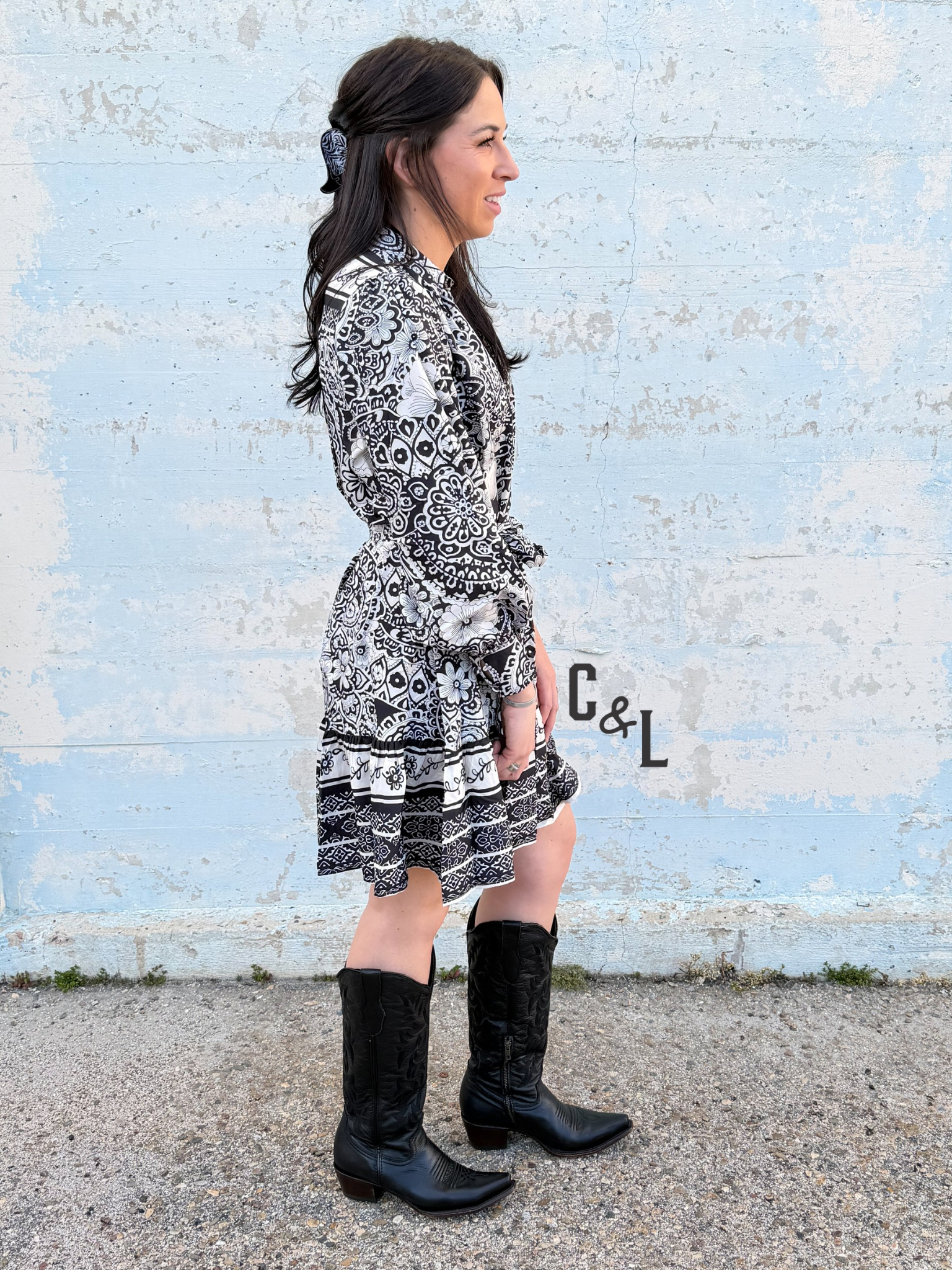 Woman wearing a black and white patterned dress and black boots standing against a textured wall.