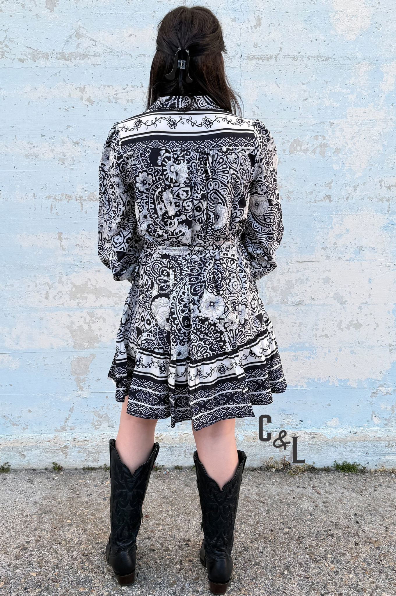 Person wearing a black and white patterned dress with cowboy boots standing against a light blue brick wall.