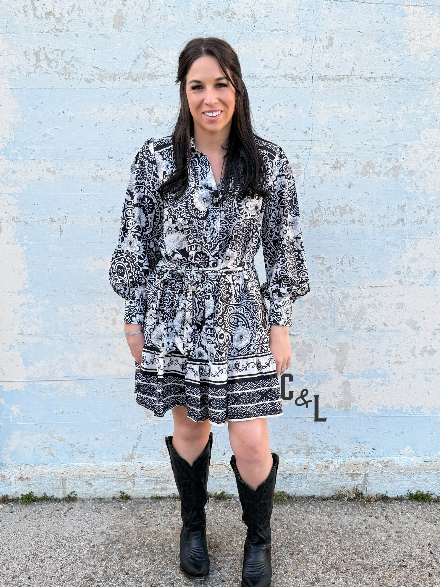 Woman wearing a black and white patterned dress with black boots standing against a light blue brick wall.