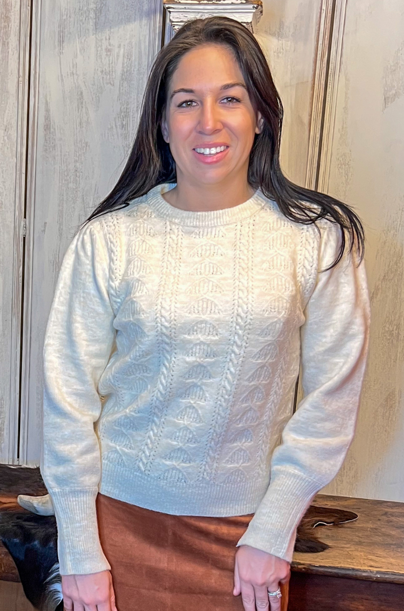 Woman wearing a light sweater and brown skirt standing in a room with wooden furniture.