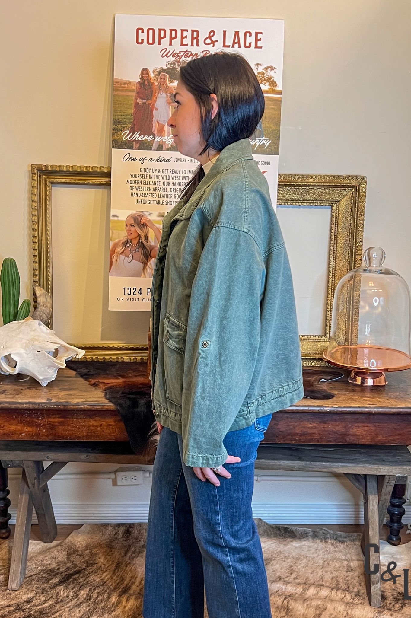 Person wearing a green jacket and blue jeans standing in front of a table with decorative items and posters.