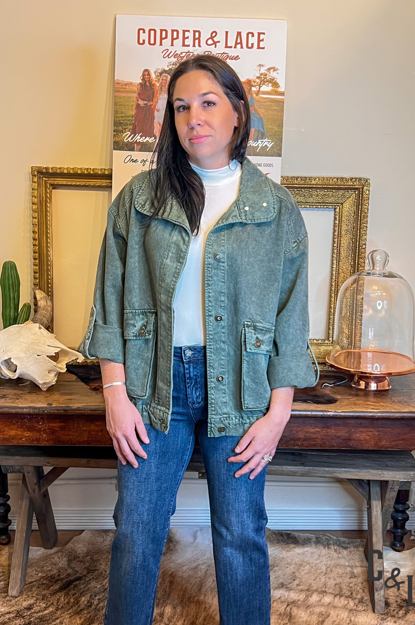 Woman wearing a green jacket and blue jeans standing in front of a table with decorative items.