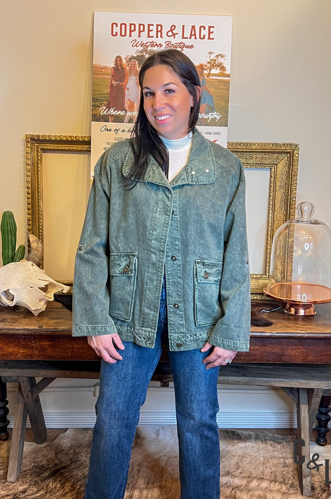 Woman wearing a green jacket denim bomber jacket and blue jeans standing in front of a 'Copper & Lace' sign.