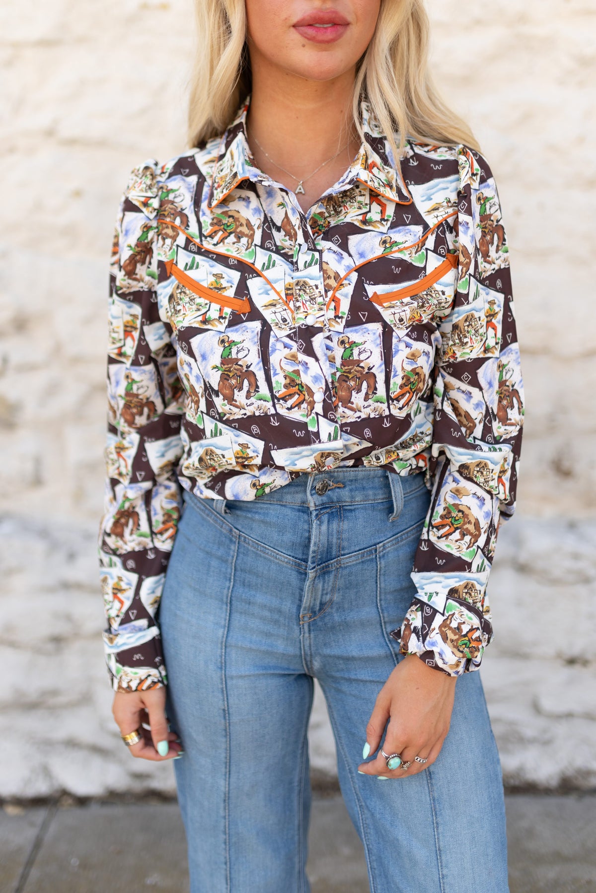 A woman wearing a long-sleeve button-up shirt with a busy collage print of vintage candy packages in brown, white, orange, and green, paired with high-waisted blue jeans, standing against a light-colored brick wall.
