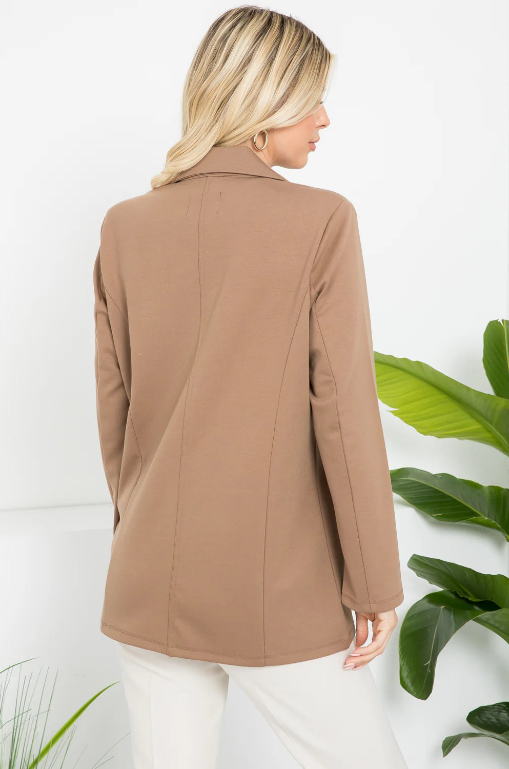 Woman wearing a brown coat with a white background and green plants.