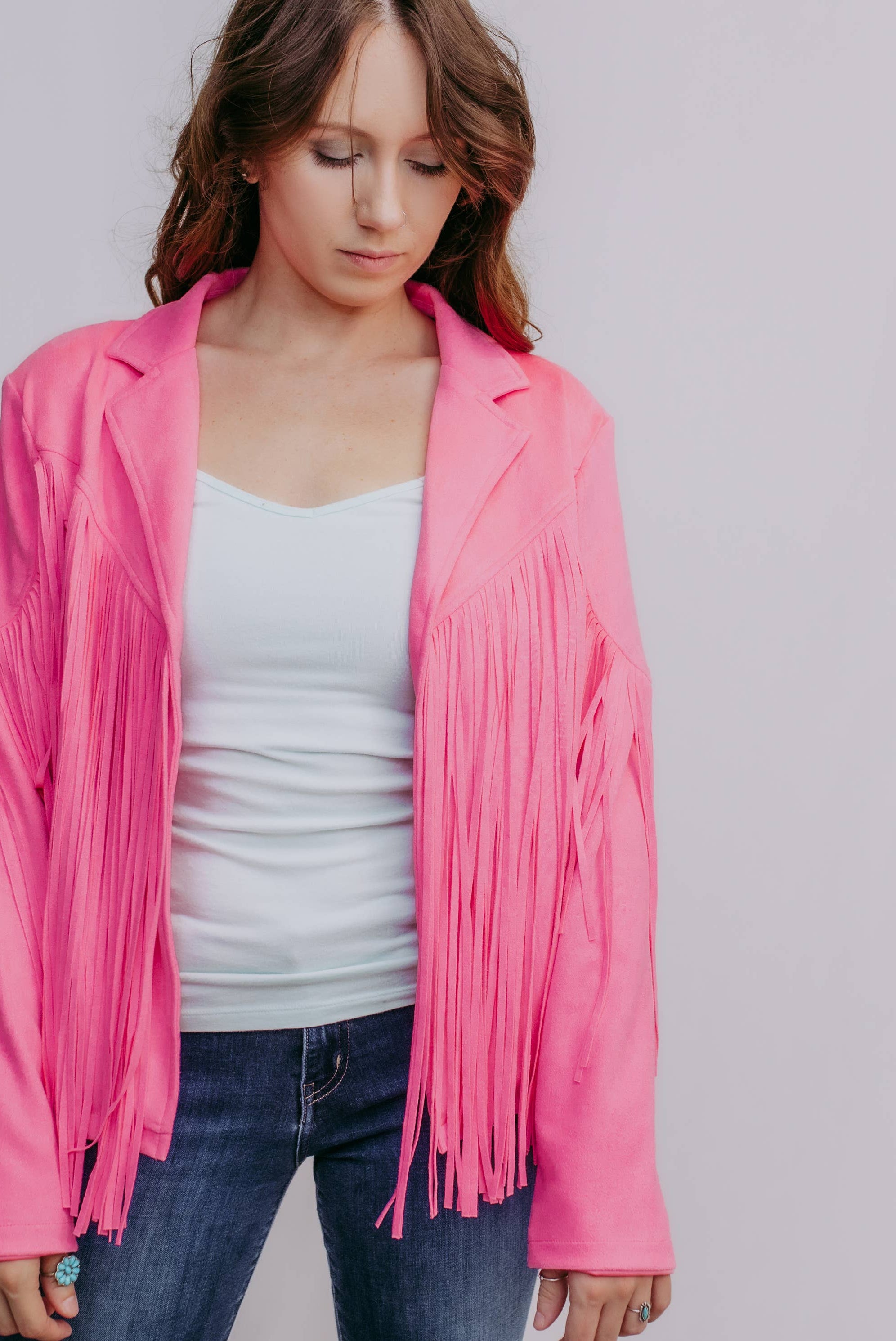 Woman wearing a pink fringed jacket over a white top and blue jeans on a plain background