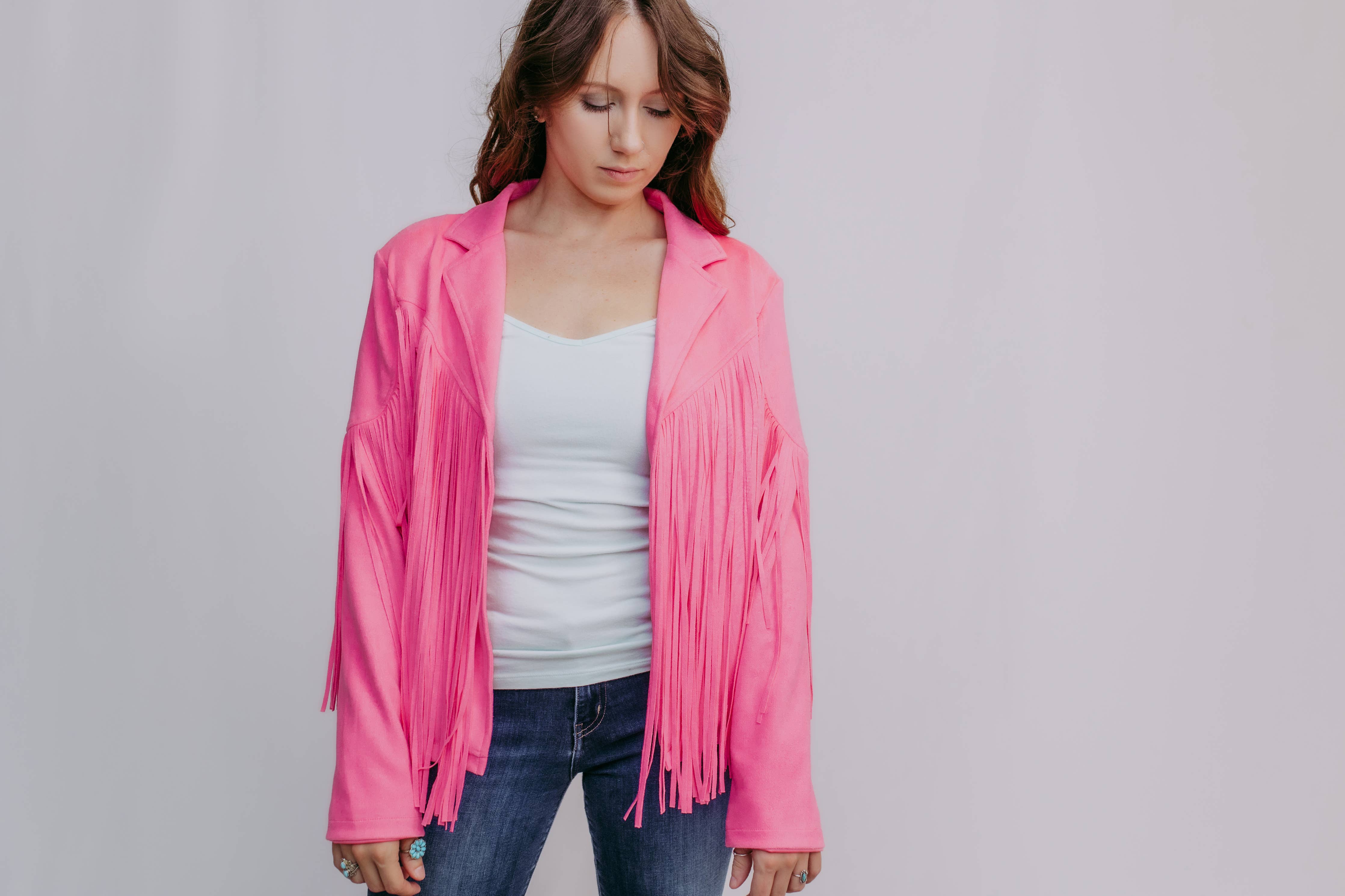 Woman wearing a pink fringed jacket over a white top and blue jeans on a plain background
