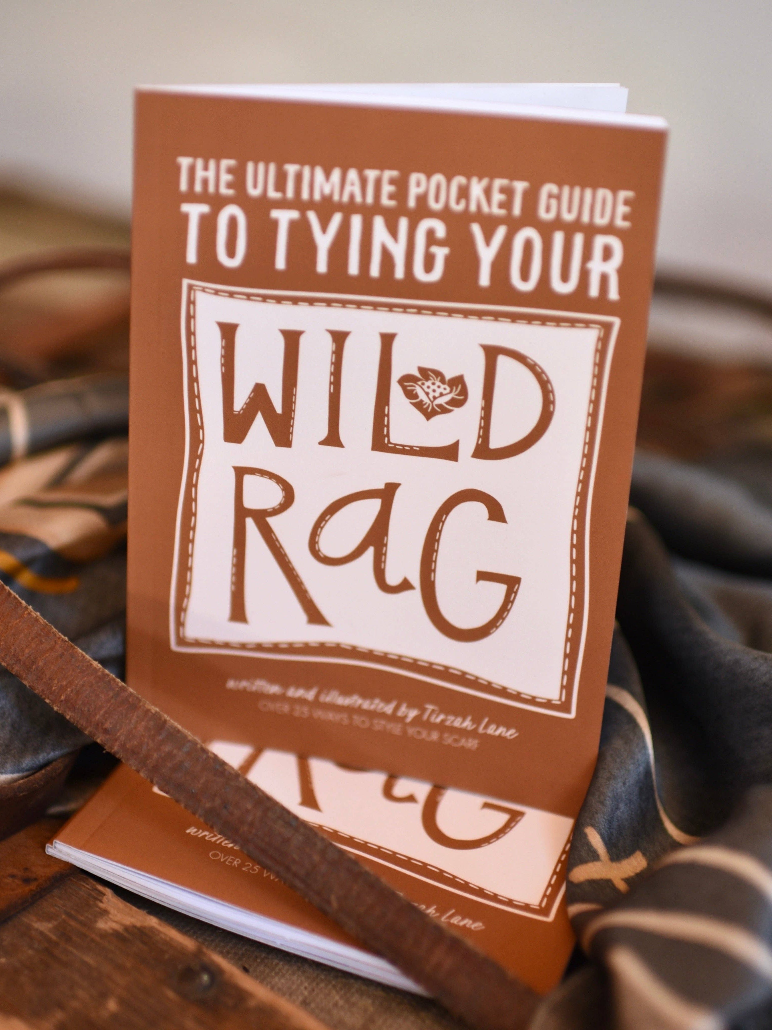 Book titled 'The Ultimate Pocket Guide to Tying Your Wild Rag' on a wooden surface with leather straps.