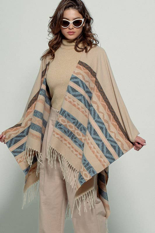 Woman wearing a beige poncho with a Tan background and light blue, brown, light rust, and grey aztec pattern design in stripes with tassel trim along bottom hem. Light weight fabric